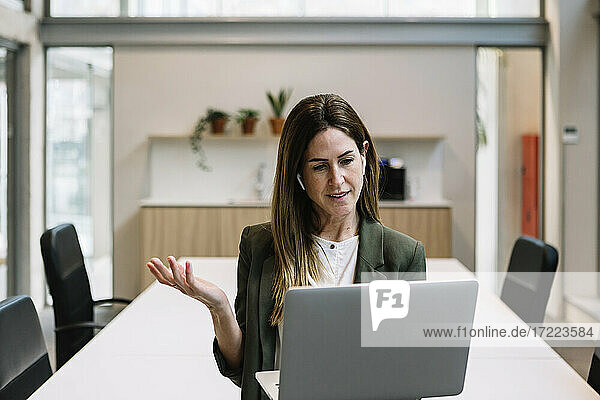 Geschäftsfrau mit In-Ear-Kopfhörern gestikuliert während eines Videogesprächs am Laptop im Büro