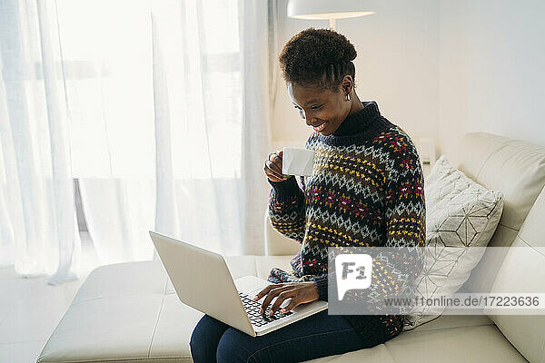 Smiling businesswoman having coffee while using laptop on sofa in living room
