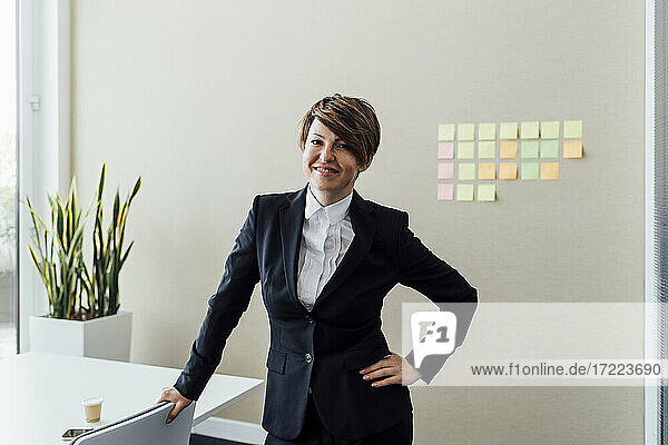 Lächelnde Unternehmerin mit Hand in der Hüfte am Schreibtisch stehend im Büro