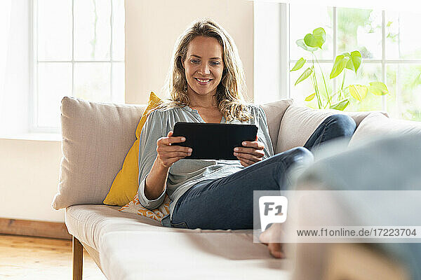 Zufriedene Frau schaut auf ein digitales Tablet  während sie auf dem Sofa im Wohnzimmer sitzt