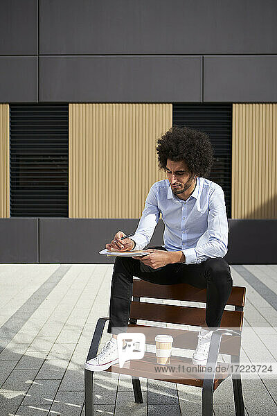 Mann mit Kaffeetasse  der auf einem Stuhl sitzend in ein Buch schreibt  während er an einem sonnigen Tag sitzt
