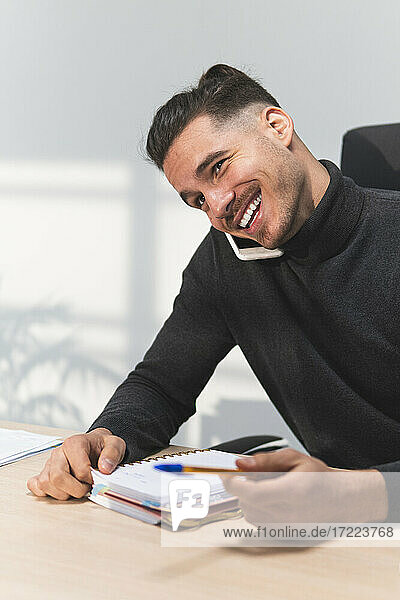 Lächelnder männlicher Finanzberater mit Tagebuch und Stift im Gespräch mit einem Smartphone im Büro