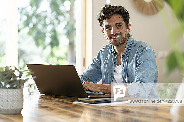 Smiling male freelancer looking away while sitting with laptop at home office