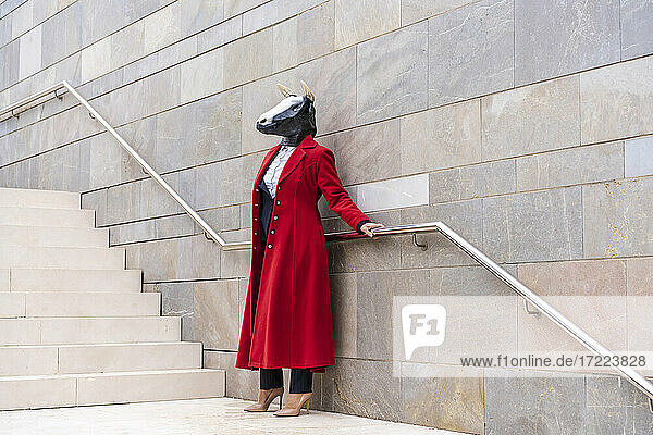 Frau mit rotem Mantel und Stiermaske hält sich an einem Geländer fest  während sie in der Nähe einer Treppe steht