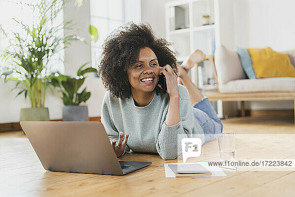 Lächelnde junge Frau  die mit ihrem Handy spricht  während sie zu Hause am Laptop liegt