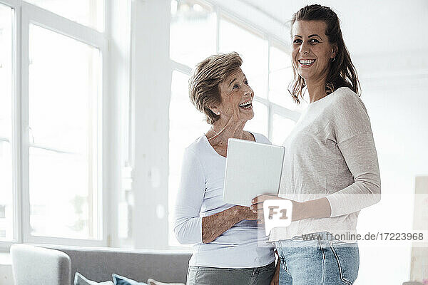 Fröhliche ältere Frau schaut auf ihre Enkelin  die zu Hause ein digitales Tablet hält