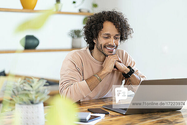 Smiling man with hands clasped looking at laptop on table at home