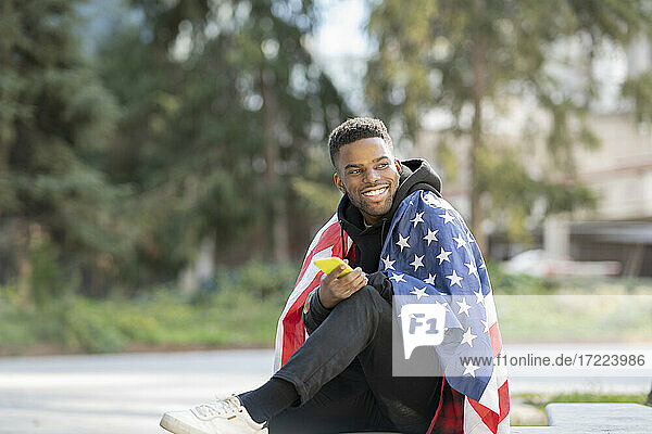 Junger Mann  eingewickelt in die amerikanische Flagge  während er im Park wegschaut