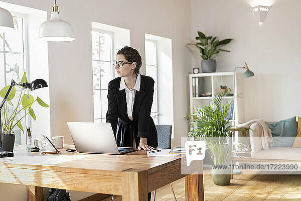 Weibliche Fachkraft  die wegschaut  während sie sich auf den Schreibtisch im Heimbüro stützt