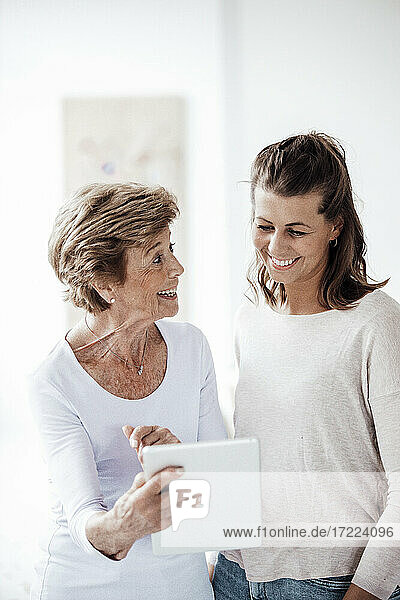 Excited senior woman holding digital tablet while talking with granddaughter at home