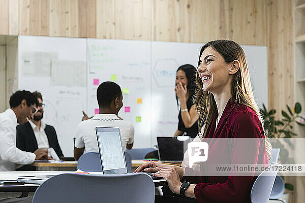 Fröhlicher Unternehmer am Laptop sitzend mit Kollegen im Hintergrund im Büro