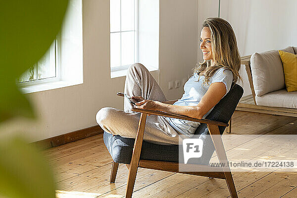 Lächelnde blonde Frau  die wegschaut  während sie mit einem digitalen Tablet auf einem Sessel zu Hause sitzt