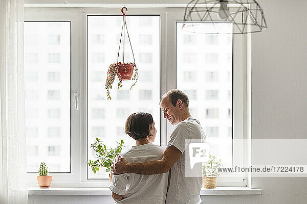 Mann und Frau schauen sich an  während sie sich in der Nähe des Fensters zu Hause umarmen