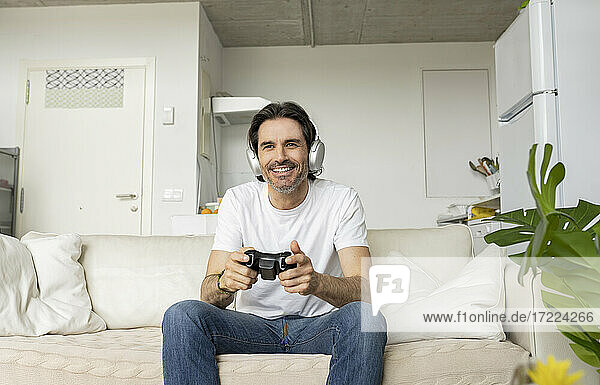 Mature man with headphones playing video game while sitting on sofa in living room