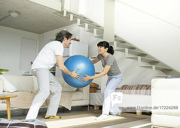 Verspieltes reifes Paar  das einen Fitnessball hält  während es im Wohnzimmer zu Hause steht