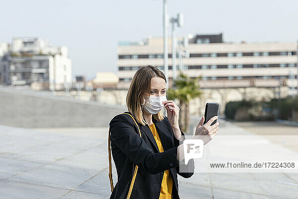 Geschäftsfrau nimmt Gesichtsmaske ab  während sie auf dem Gehweg ein Selfie mit dem Handy macht