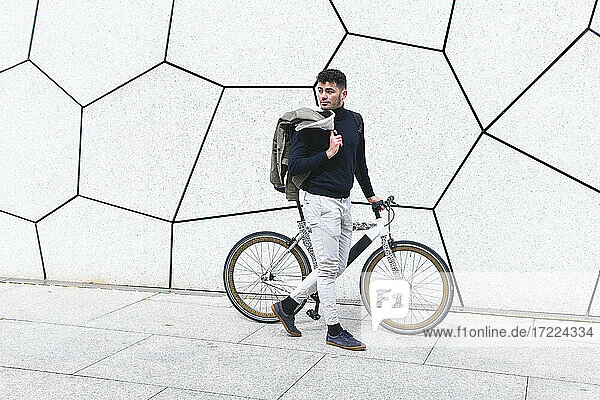 Young man looking away while walking with bicycle on footpath