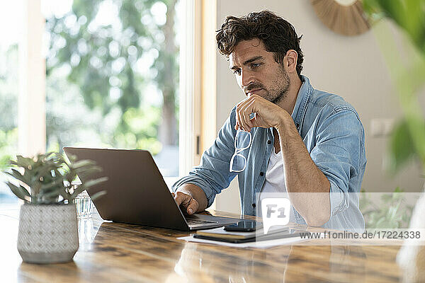 Concentrated male freelancer holding eyeglasses while working on laptop at table