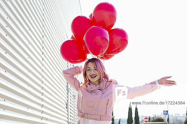 Glückliche Frau mit Luftballons an einer Wellblechwand an einem sonnigen Tag