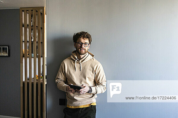 Man wearing eyeglasses holding digital tablet while leaning on wall at home