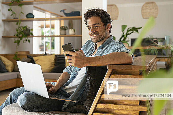 Smiling male freelance worker with laptop using smart phone while sitting couch in living room