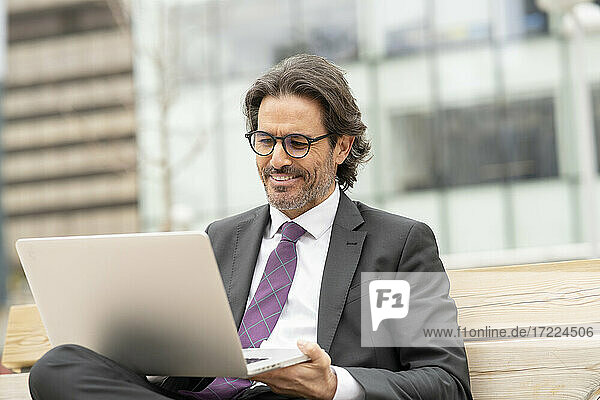 Zufriedener Geschäftsmann mit Brille  der einen Laptop benutzt  während er auf einer Bank sitzt