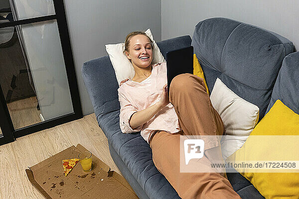 Woman using digital tablet while resting by food on sofa at home