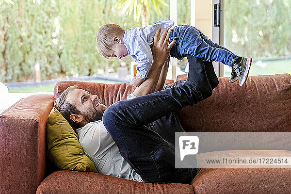 Lächelnder Vater hebt seinen Sohn hoch  während er auf dem Sofa im Wohnzimmer liegt