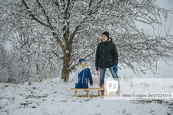 Father and son standing with sled in front of tree during winter