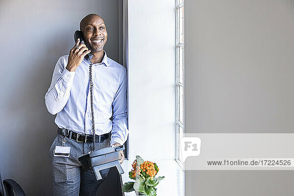 Smiling male entrepreneur talking on landline phone while leaning on wall at office