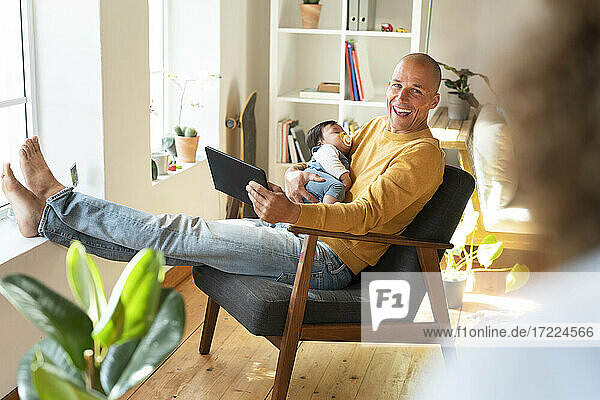 Smiling father with digital tablet and sleeping baby looking at woman in living room