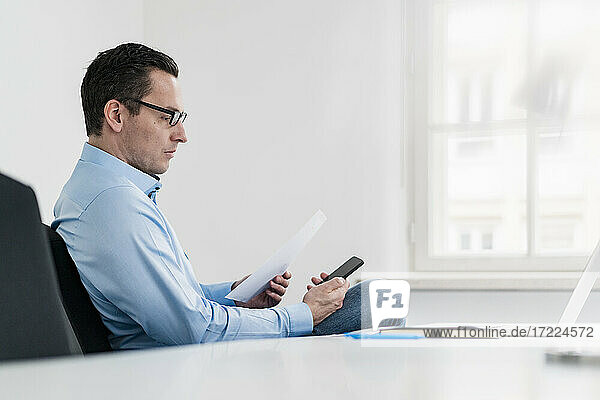 Geschäftsmann  der ein Dokument hält  während er ein Smartphone im Büro benutzt