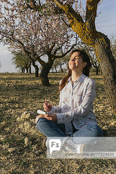 Lächelnde Frau sitzt mit Notizblock unter einem Mandelbaum