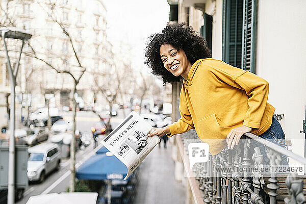Fröhliche junge Frau mit Zeitung am Balkongeländer lehnend