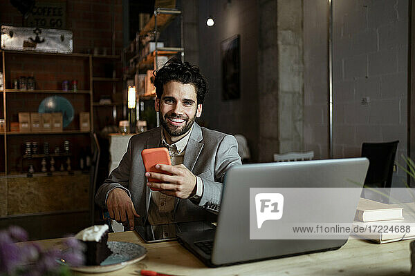 Glücklicher Geschäftsmann mit Laptop und Mobiltelefon in einem Café sitzend