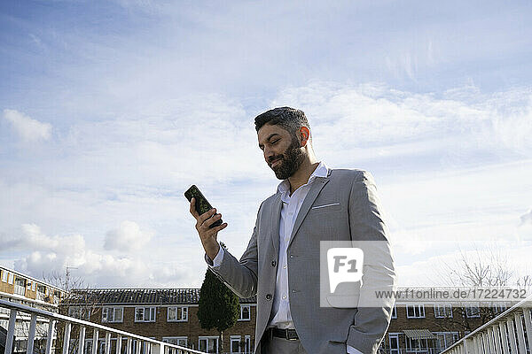 Lächelnder Geschäftsmann  der ein Mobiltelefon vor dem Himmel benutzt