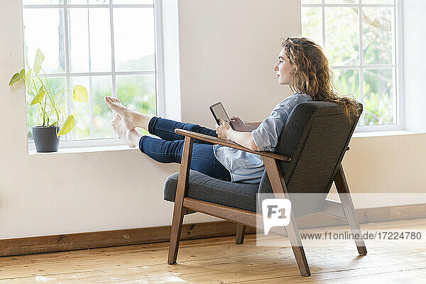 Junge Frau hält digitales Tablet  während sie zu Hause auf einem Sessel sitzt