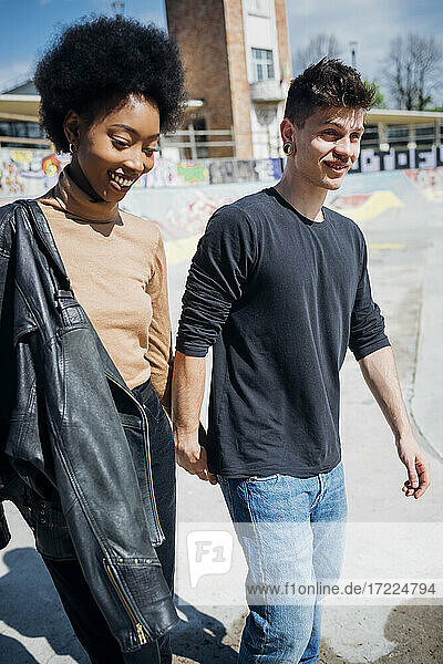 Smiling multi ethnic couple walking while holding hands at skateboard park during sunny day