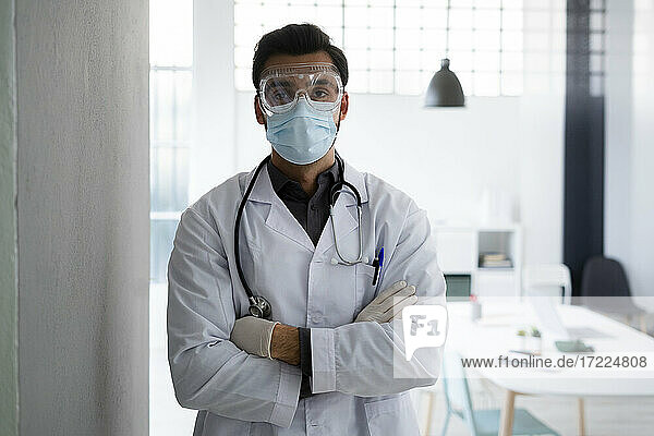 Männlicher Mitarbeiter im Gesundheitswesen mit Gesichtsschutz  stehend mit verschränkten Armen im Krankenhaus