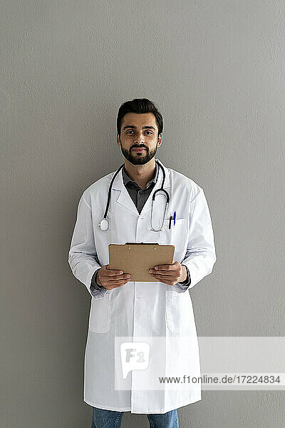 Männlicher Arzt mit Klemmbrett vor einer Wand stehend