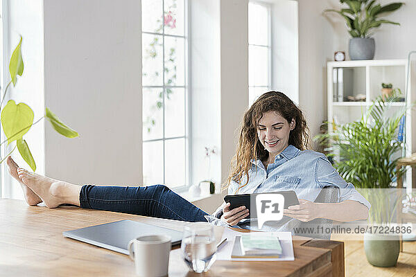 Lächelnde Unternehmerin  die auf ein digitales Tablet schaut  während sie mit hochgelegten Füßen am Schreibtisch sitzt