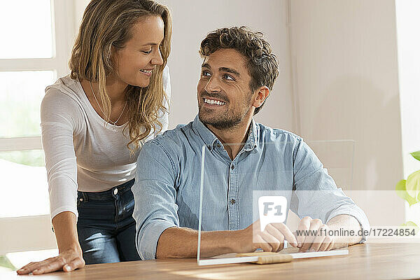 Glückliches Paar  das sich gegenseitig ansieht  während es mit einem transparenten Bildschirm zu Hause sitzt