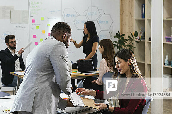Männliche und weibliche Unternehmer diskutieren über Dokumente mit Kollegen im Hintergrund im Büro