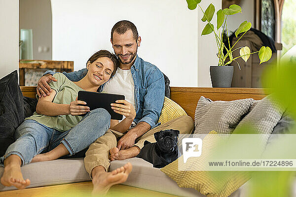 Man embracing woman holding digital tablet while sitting with Pug dog on sofa at home