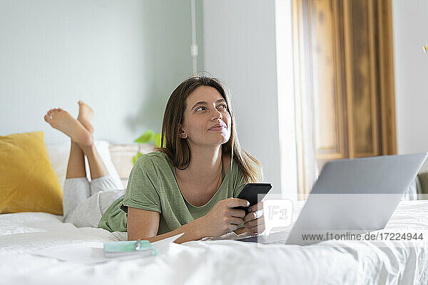 Thoughtful young woman with smart phone lying in front of laptop on bed at home