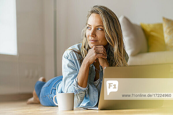 Thoughtful woman with hand on chin looking away while lying on floor in front of laptop