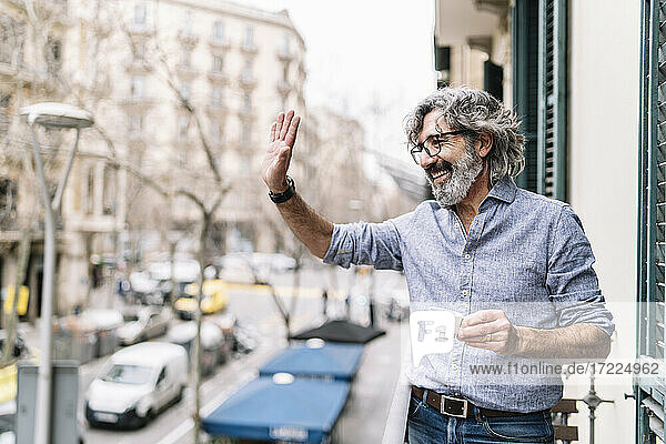 Fröhlicher älterer Mann winkt mit der Hand  während er auf dem Balkon eines Hauses Kaffee trinkt