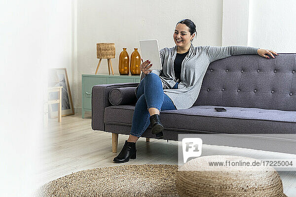 Lächelnde Frau  die ein digitales Tablet benutzt  während sie auf dem Sofa im Wohnzimmer sitzt