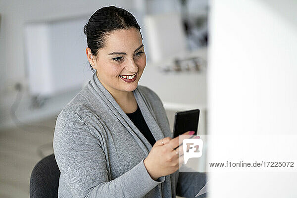 Lächelnde weibliche Fachkraft  die im Büro zu Hause ein Mobiltelefon benutzt