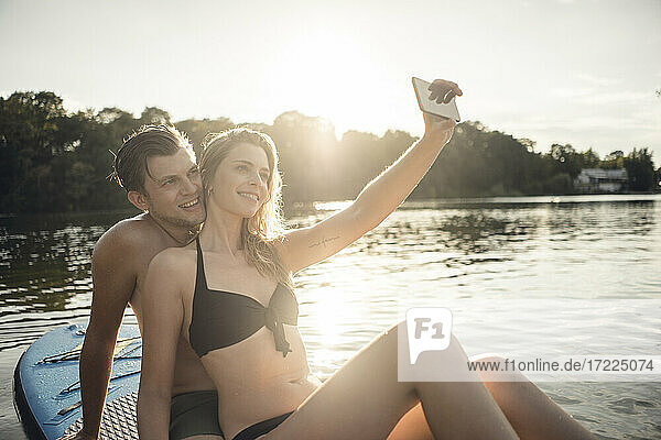 Happy couple sitting on paddleboard on a lake  taking smartphone selfies
